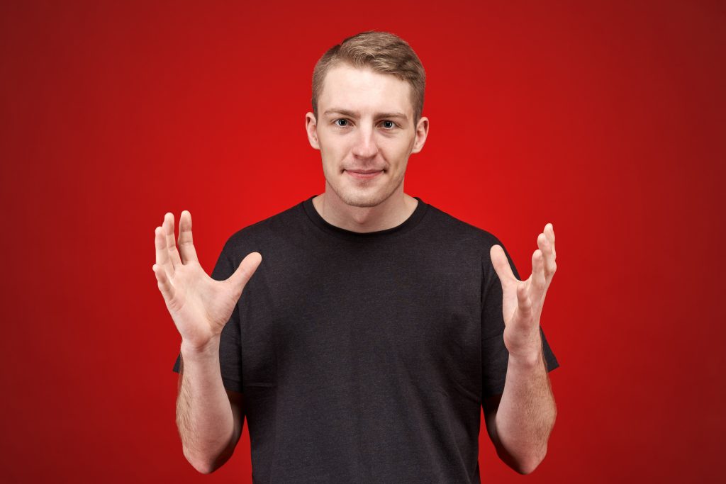 gestures - Serious angry young male model dressed in a black t-shirt on a red background, holding two hands up in a sign of indignation, discontent frowning face and trying to sink in the bad news